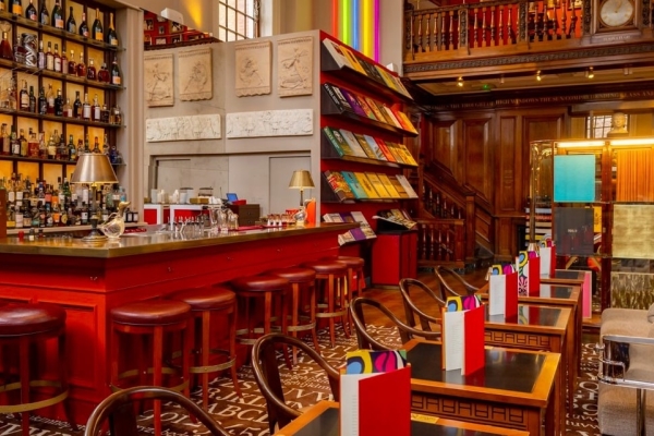 The interior of the Maison Assouline, with tables and chairs, menus, a bar with stools around it and lamps on top, and shelves full of bottles and books in the background